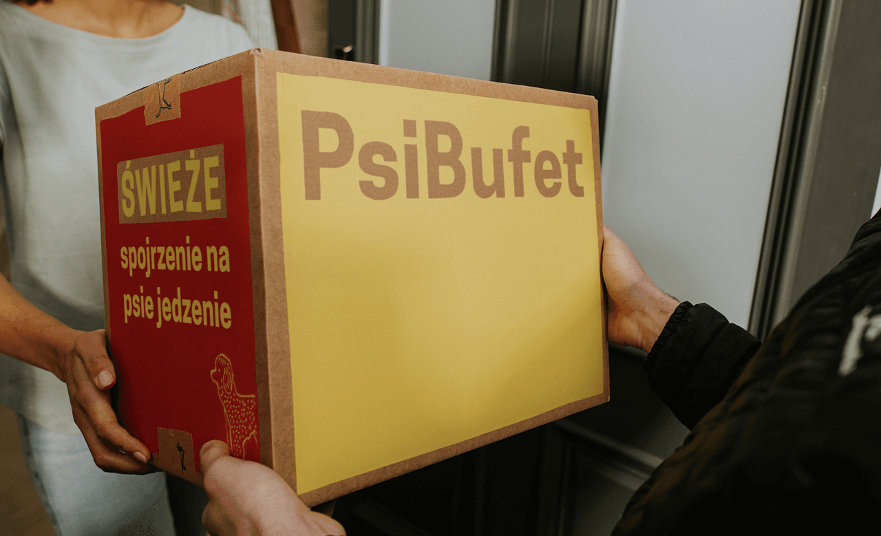 Two people exchanging a large box labeled "PsiBufet" with Polish text, indoors.