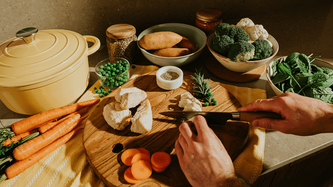 Hands slicing cooked chicken on a wooden cutting board surrounded by carrots, peas, sweet potatoes, broccoli, cauliflower, and spinach.