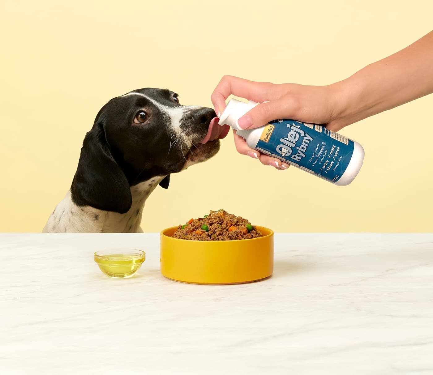 A black-and-white dog licks a bottle held by a person near a bowl of dog food and a small dish of oil on a marble surface.