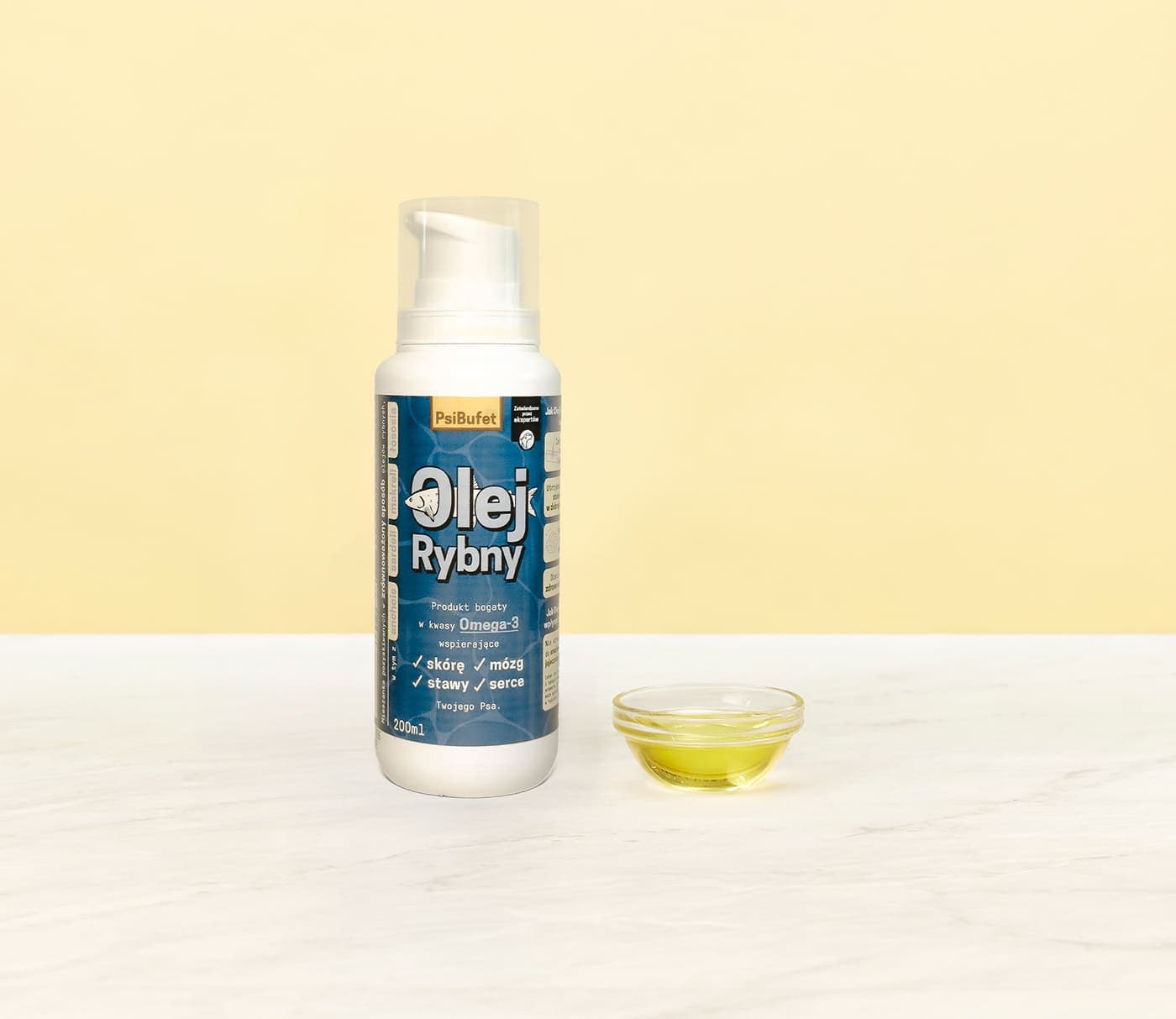 A bottle of "Olej Rybny" fish oil next to a small glass dish with oil, placed on a white marble surface against a light yellow background.
