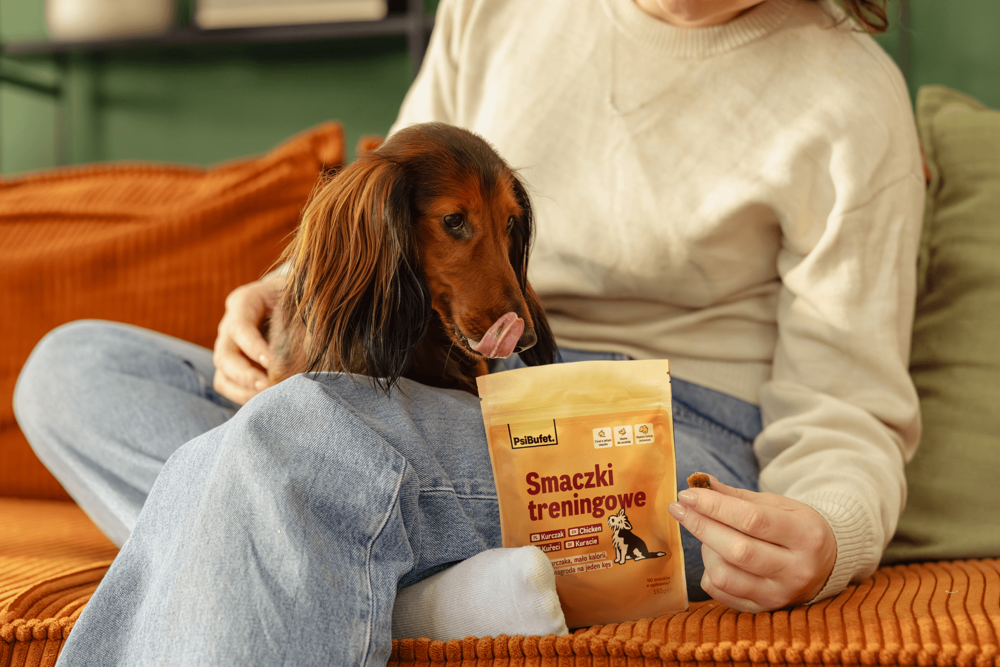 A dachshund dog sits on a person's lap, eagerly licking its lips while looking at a package of PsiBufet training treats held in the person's hand.