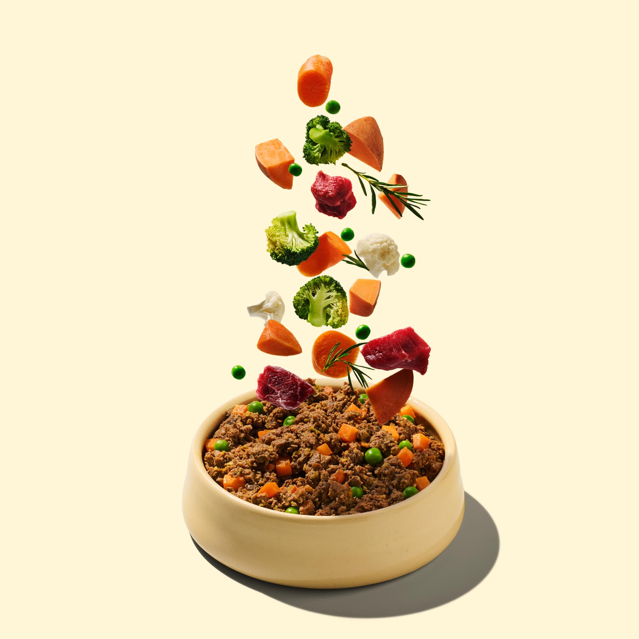 A bowl of pet food with various ingredients like meat, carrots, broccoli, peas, and herbs levitating above against a light beige background.