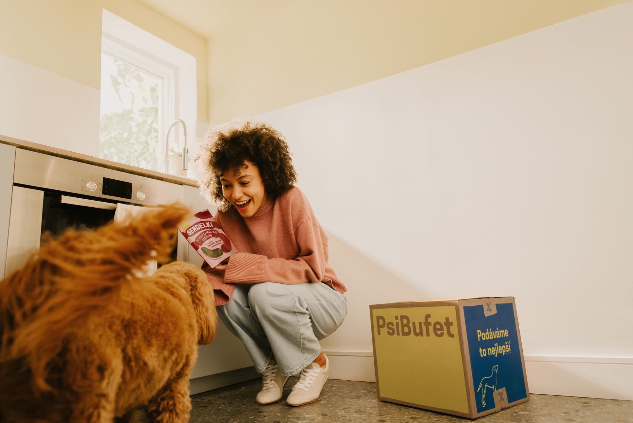 Woman standing next to the PsiBufet box in the kitchen feeding Serdelki a cavapoo dog
